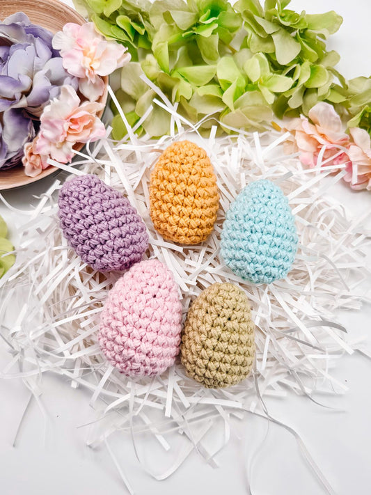 Five crocheted eggs in purple, orange, blue, pink, and green rest on white shredded paper, surrounded by pink and purple flowers with green leaves—a perfect handmade gift to accompany your unique jewellery collection.