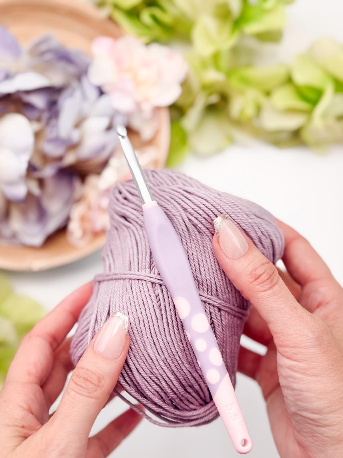Hands holding a lavender-colored yarn ball with a crochet hook, surrounded by soft-focus flowers, crafting the perfect handmade gift.