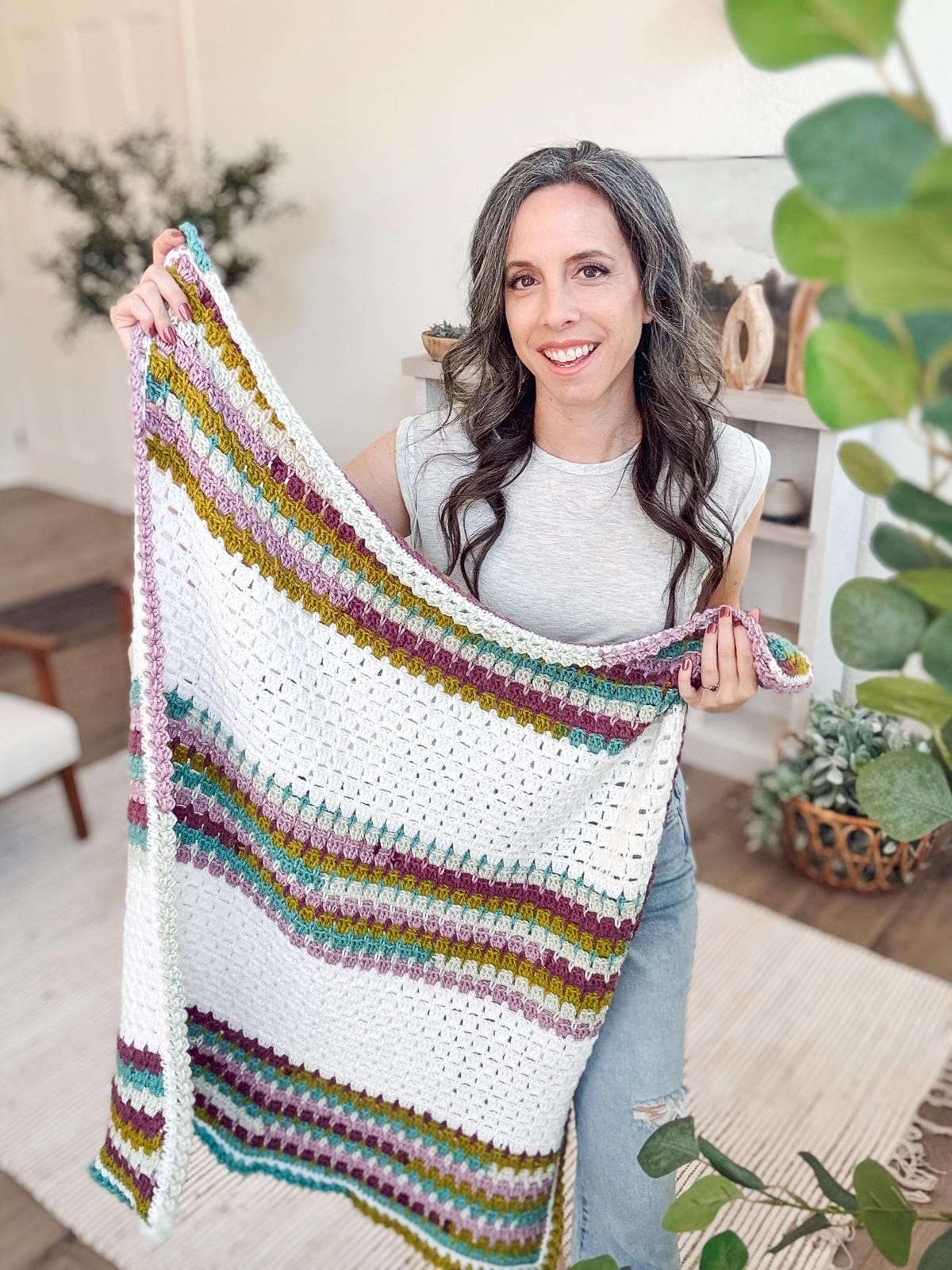 A woman holds a colorful crocheted blanket, standing in a room adorned with lush plants and eclectic decor, offering the perfect setting for showcasing unique handmade gifts.