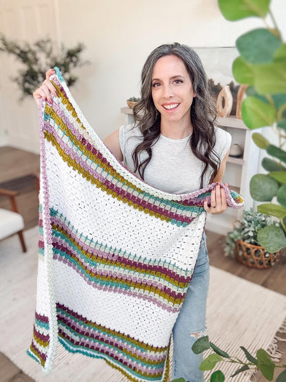 White crochet blanket with pink, mustard, and teal stripes held up by a smiling woman in a bright room with plants