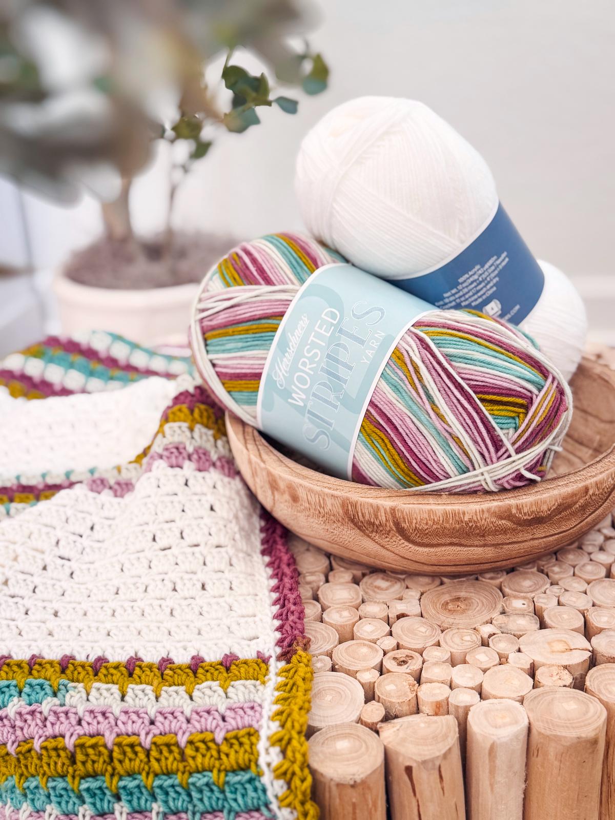 A multicolored crochet blanket draped over a white armchair next to a wooden table, offering the perfect touch of charm for your home. A plant stands in the background on a striped rug, reminiscent of an inviting space filled with handmade gifts and unique jewelry treasures.
