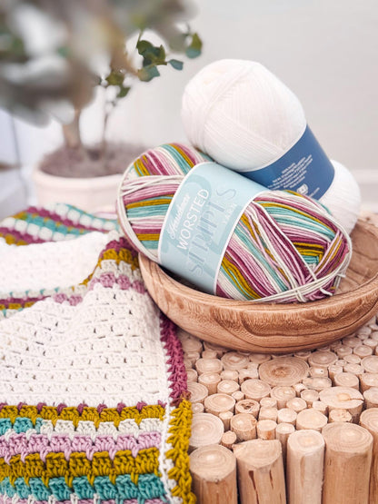Striped and white yarn skeins in a wooden bowl beside a white crochet blanket with pink, teal, and mustard stripes