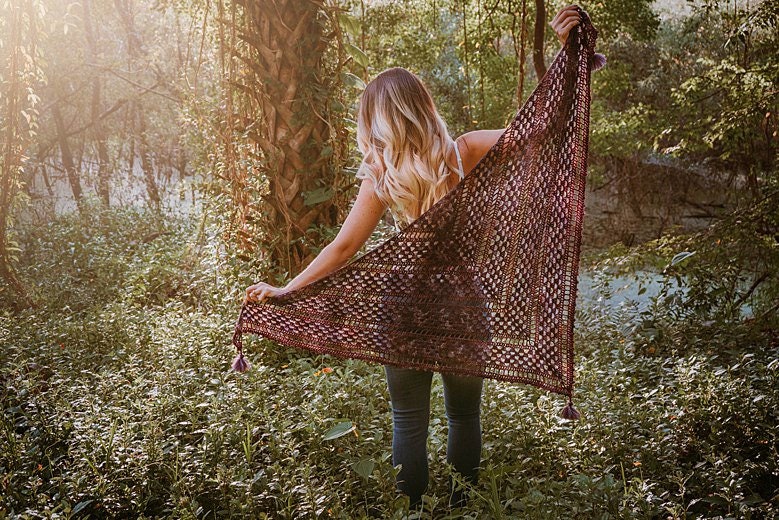 Amidst the lush greenery, a person stands holding a large triangular crochet shawl with tassels, reminiscent of vintage clothes. Sunlight filters through the trees, casting enchanting patterns around them.
