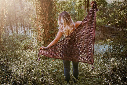 Amidst the lush greenery, a person stands holding a large triangular crochet shawl with tassels, reminiscent of vintage clothes. Sunlight filters through the trees, casting enchanting patterns around them.
