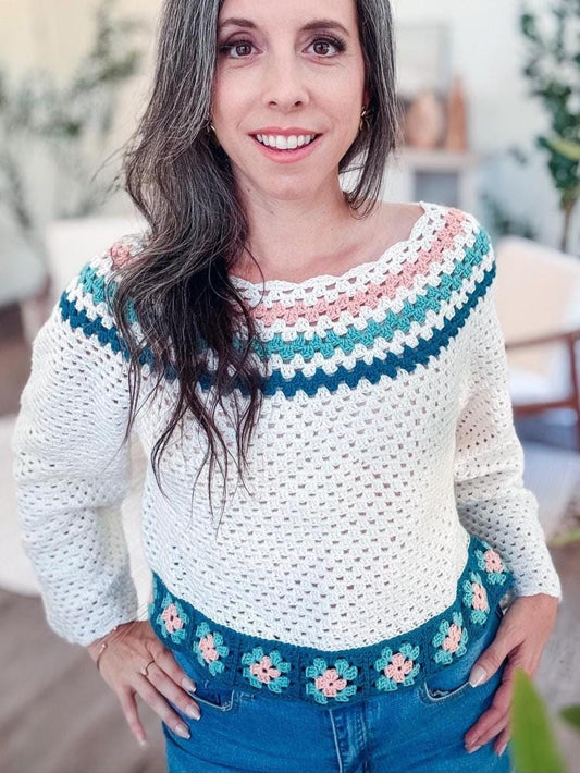 A person with long hair, adorned in a white crocheted sweater with colorful accents, stands indoors near plants and furniture, showcasing their vintage clothes.