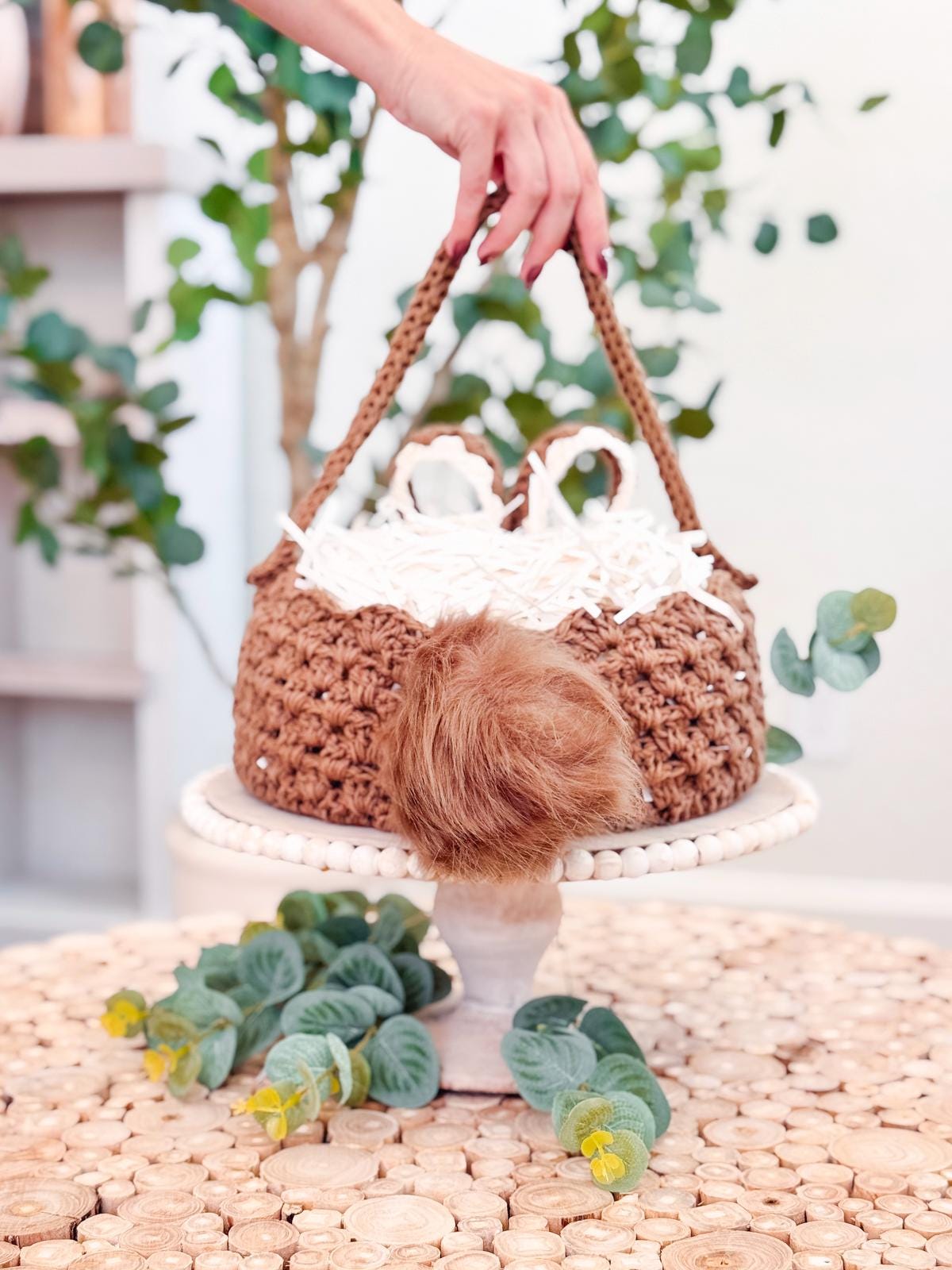 A hand holds a crocheted basket with handles on a white stand. Inside the basket, resembling a handmade gift, there is white shredded paper and a brown furry item. Green leaves lie around the base of the stand.