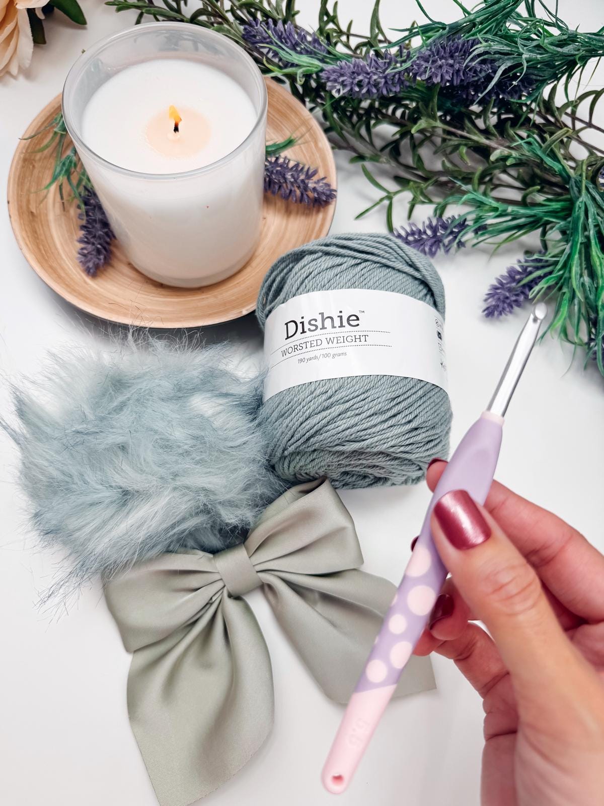 A hand holds a crochet hook near a gray yarn ball labeled "Dishie," alongside a lit candle, a gray bow, and faux lavender on a white surface, creating the perfect setting for crafting a unique handmade gift.