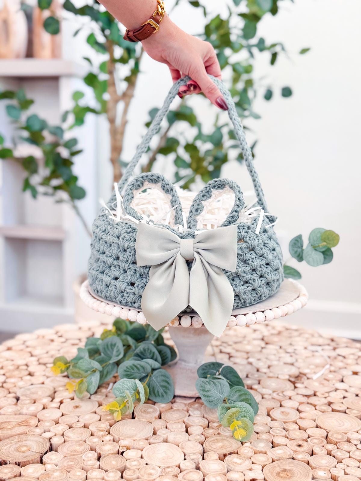 A hand holds a crochet basket with a large bow, placed on a round wooden table. The table is topped with a plant, alongside unique jewellery that catches the eye, while greenery decorates the background.