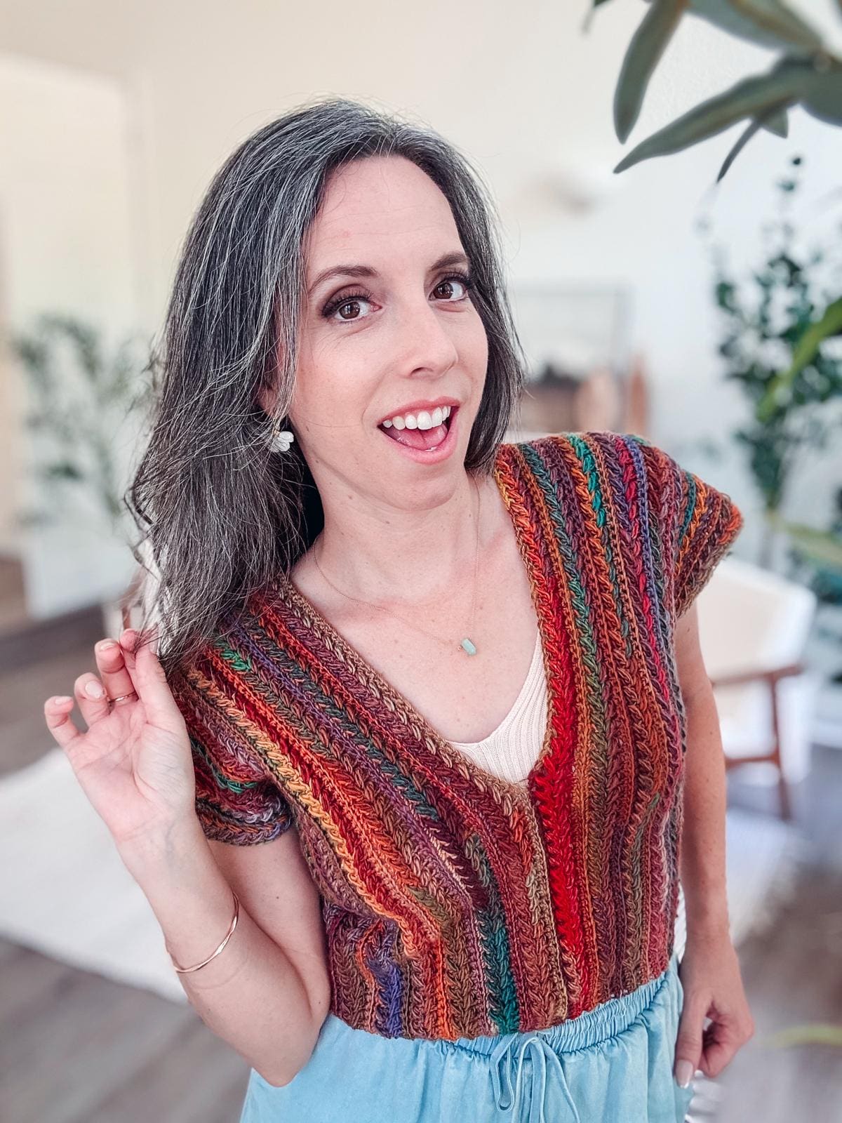 A woman with long gray hair smiles at the camera, wearing a colorful crocheted top paired with light blue pants. She stands indoors, surrounded by plants and furniture, her look accentuated by unique jewelry that adds a touch of elegance to her vintage style.