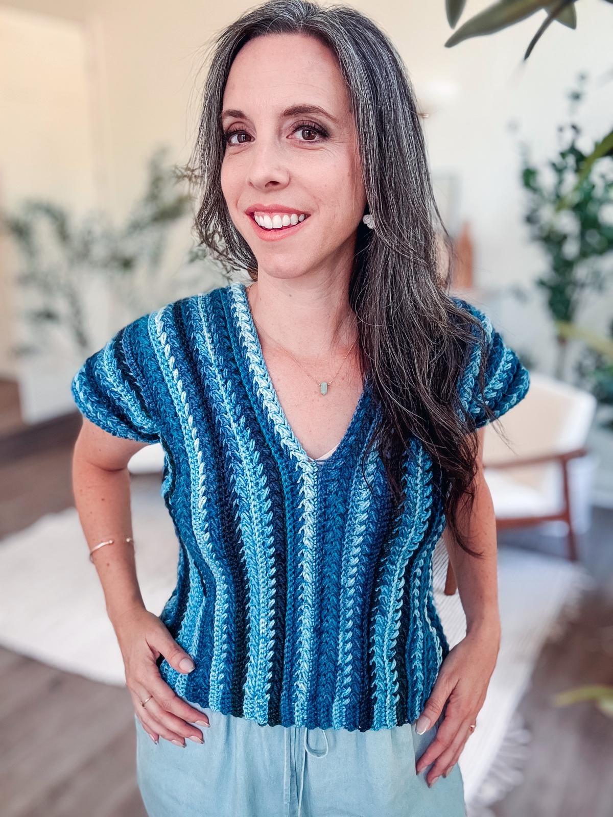 Woman with long hair in a blue striped top smiles indoors, hands on hips. The background features light walls, green plants, and a chair adorned with unique jewelry, adding a touch of elegance.