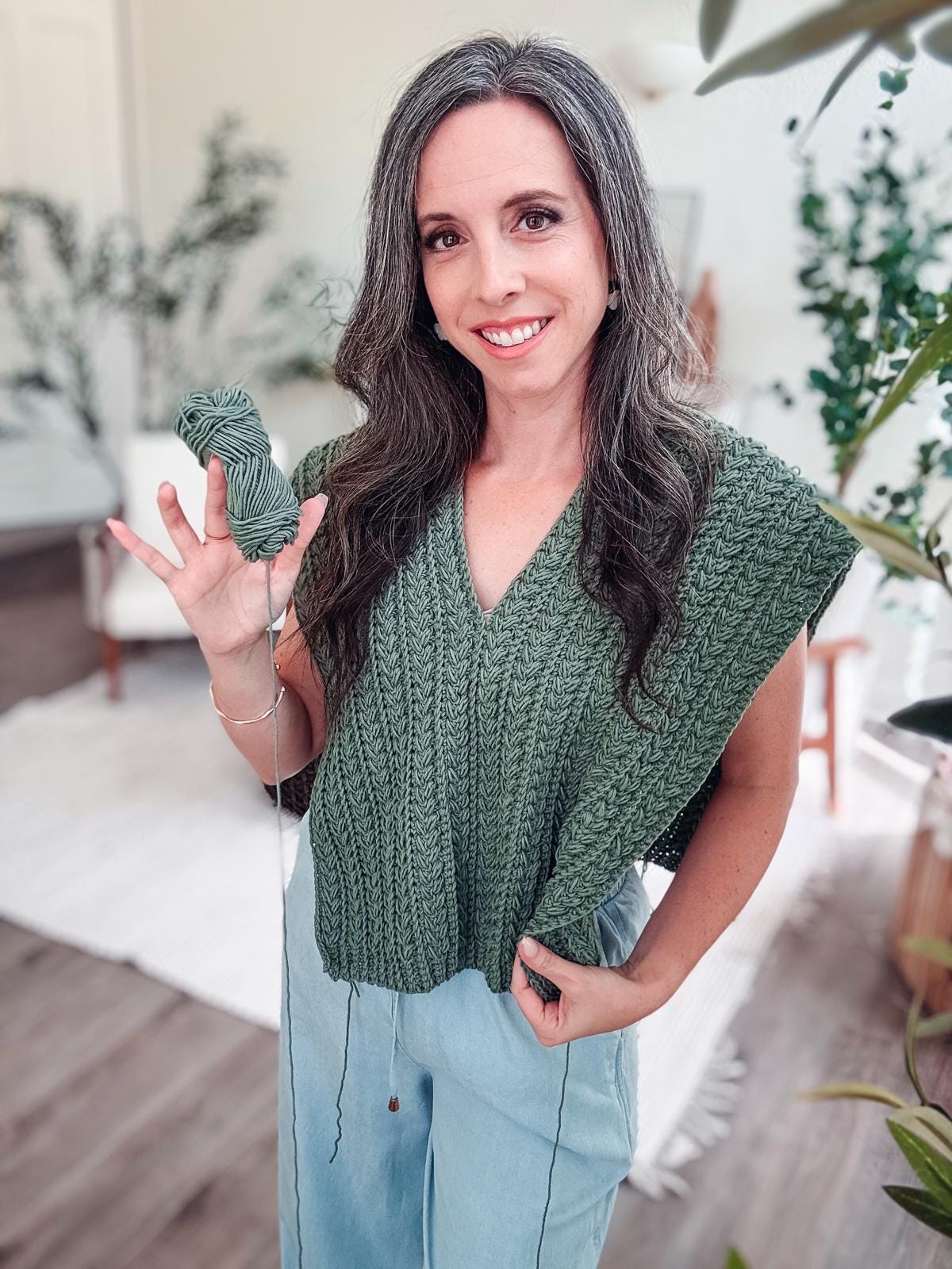 A person wearing a green crochet vest in progress, holding green yarn. They are standing indoors with plants and a rug in the background, surrounded by vintage clothes that add a nostalgic charm.