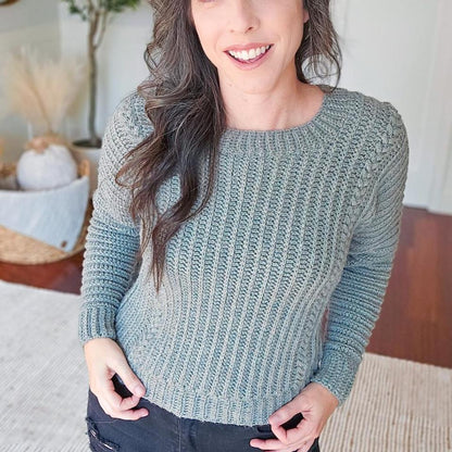 Autumn Wheat Adult Sweater crochet pattern: sage green ribbed crochet sweater with braided sleeve detail worn by a smiling woman with black jeans