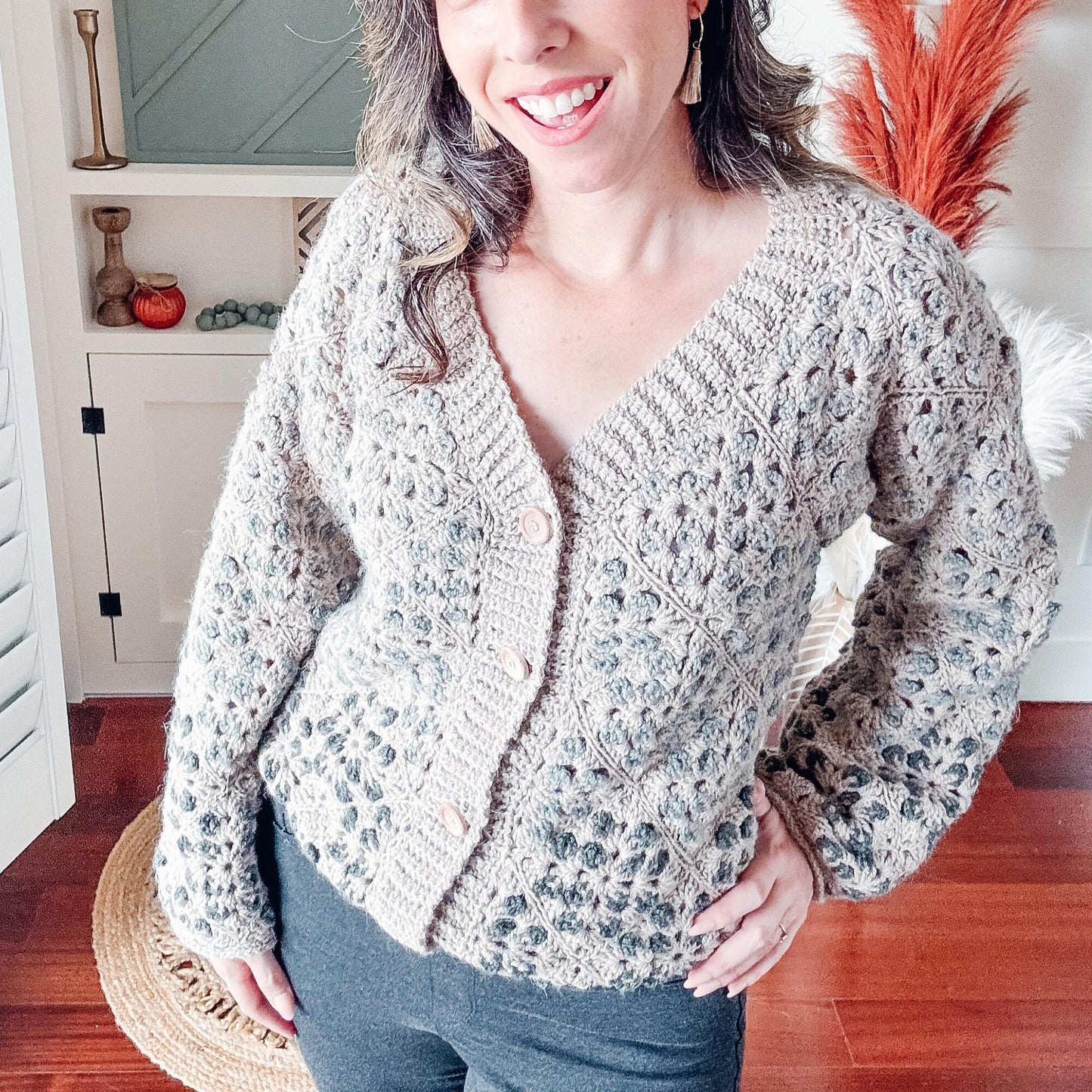 A woman in a crocheted beige cardigan and dark pants stands indoors, smiling with one hand on her hip. Decor includes shelves, vases, dried plants, and a white shuttered window adorned with unique jewellery.