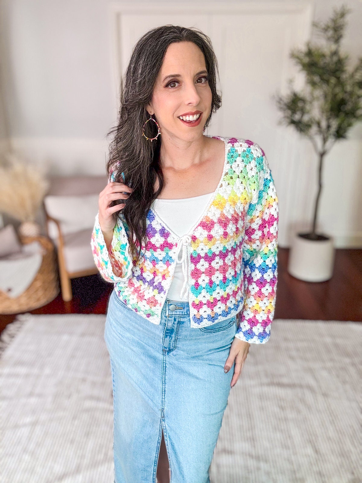 A woman smiling, wearing a colorful crocheted cardigan over a white top and a light blue denim skirt, stands indoors with a hand resting on her hip, showcasing her love for vintage clothes.