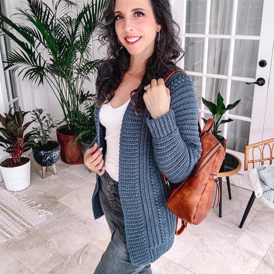 Woman with long hair, wearing a blue crochet cardigan and jeans, holds a brown backpack. She stands in a room adorned with potted plants and a glass door, exuding charm like unique jewelry complementing vintage clothes.