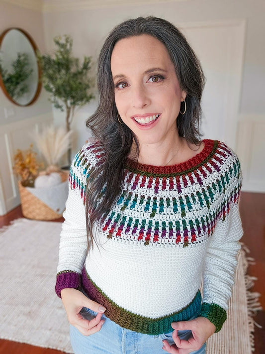 A person with long hair smiles, wearing a white crochet sweater with colorful mosaic patterns. They stand in a room with a mirror, plant, and woven basket.