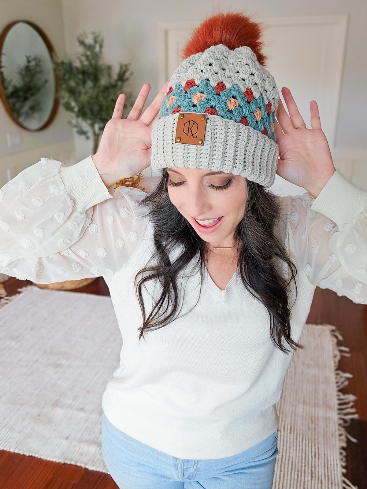 Person wearing a knitted beanie with colorful patterns and a pom-pom, raising hands near head, standing in a room with vintage clothes, surrounded by a mirror and plants.