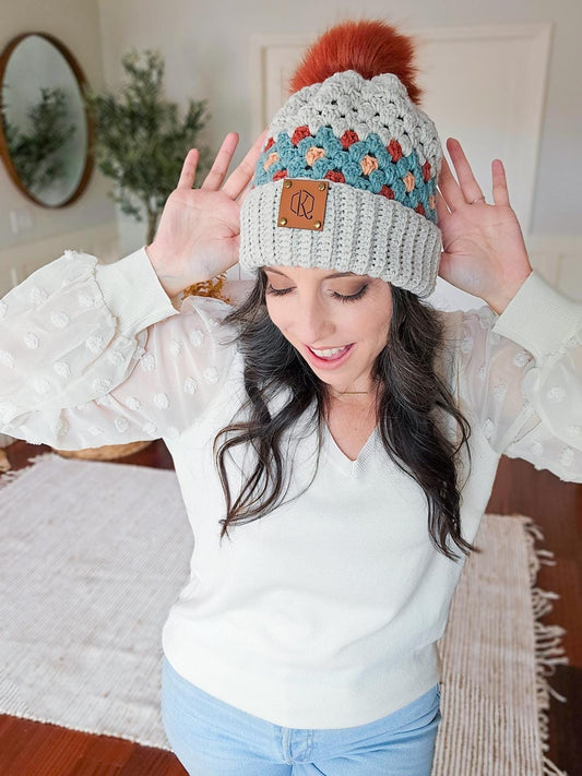 Person wearing a knitted beanie with colorful patterns and a pom-pom, raising hands near head, standing in a room with vintage clothes, surrounded by a mirror and plants.