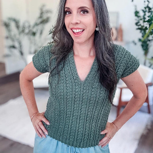 A woman with long gray hair smiles while standing indoors, wearing a green crochet top and blue pants, accessorized with unique jewellery.