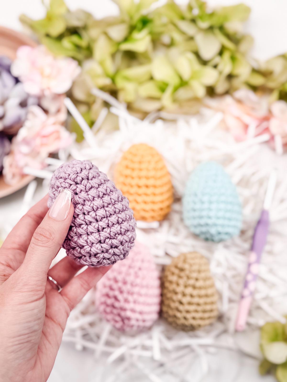 A hand holds a purple crocheted egg above a surface adorned with four other vibrant eggs and decorative flowers, offering a charming handmade gift that captures the essence of artistry and craft.