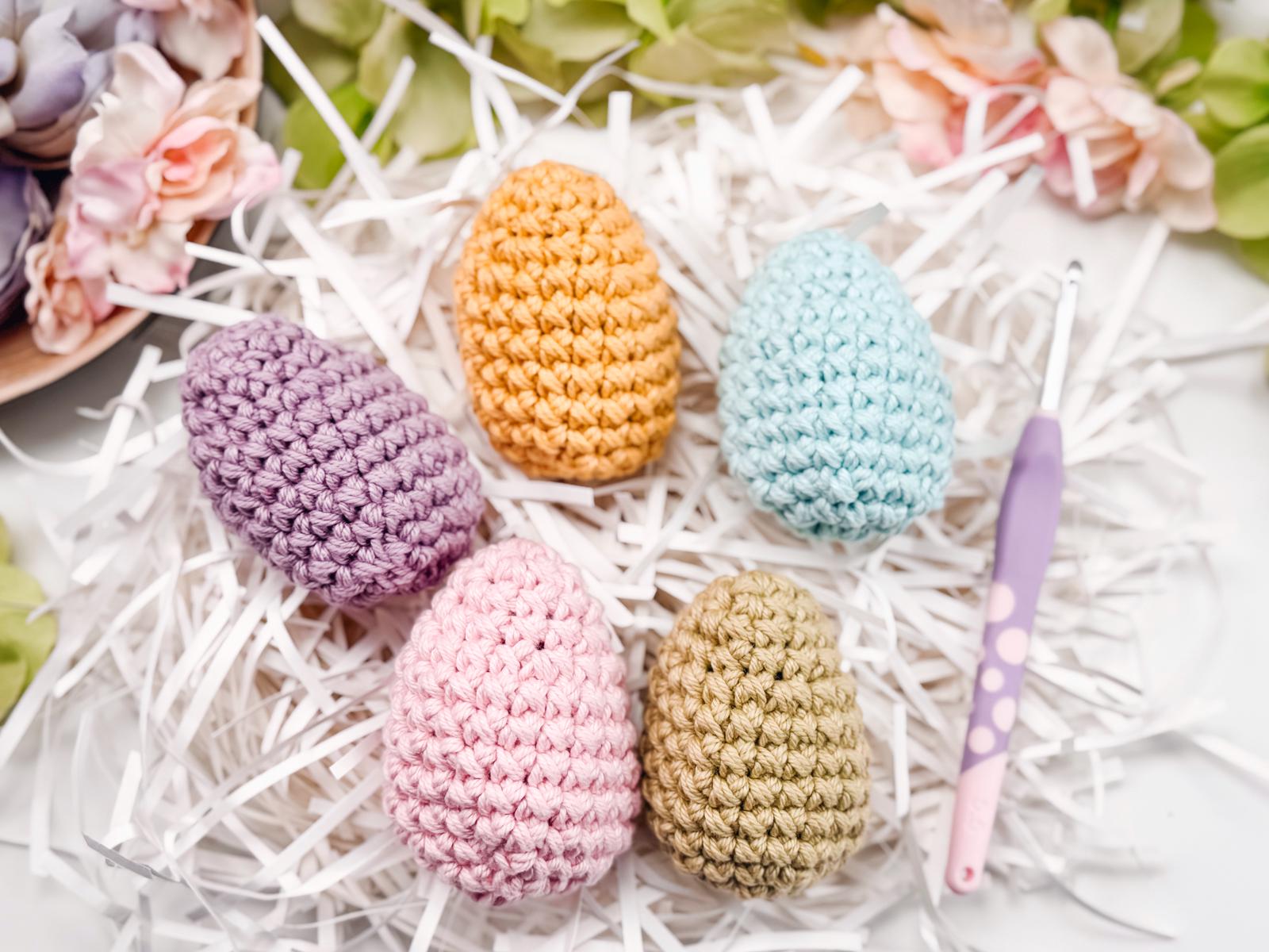 Crocheted eggs in pastel colors rest on white shredded paper, surrounded by pink and green flowers. A crochet hook lies beside them, offering a delightful handmade gift perfect for any occasion.