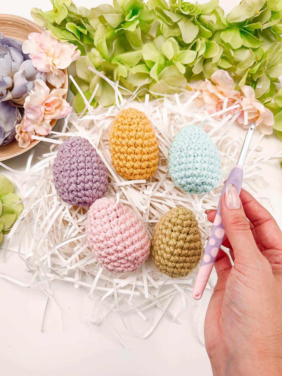 Five colorful crocheted eggs rest on a nest of white shredded paper, surrounded by flowers, with a hand skillfully holding a crochet hook, transforming this scene into the perfect handmade gift.