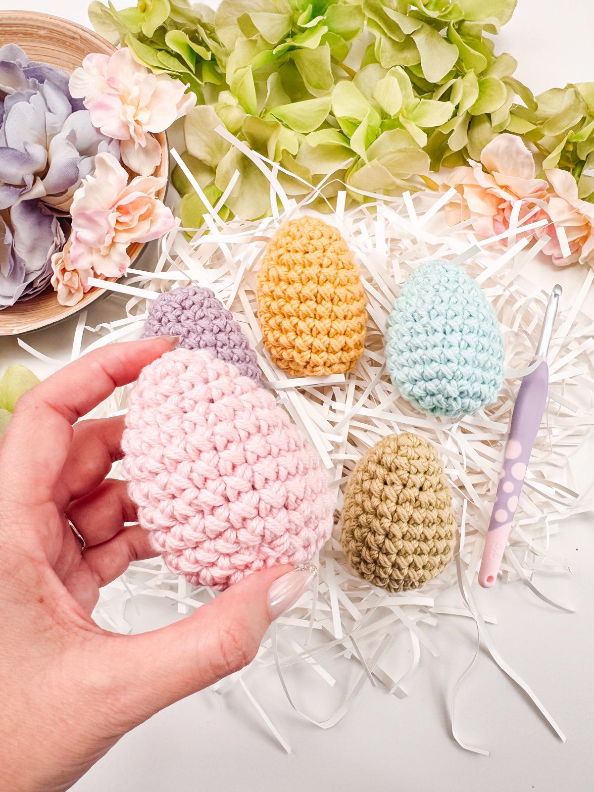 A hand holds a pink crocheted egg above a nest of white paper, surrounded by other colorful crocheted eggs, pastel flowers, and a crochet hook—an exquisite handmade gift that adds charm to any vintage clothes display.