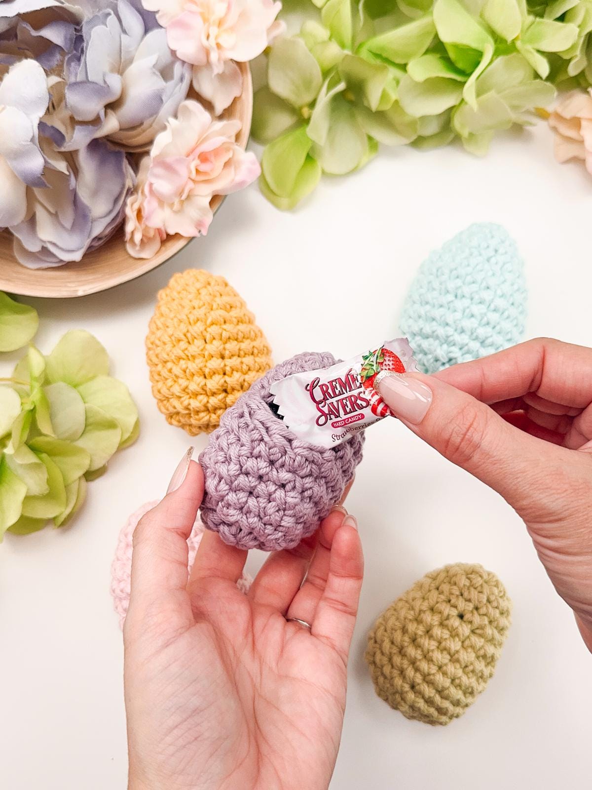 Hands placing a candy inside a crocheted egg, surrounded by pastel-colored crochet eggs and artificial flowers on a table, creating the perfect setting for presenting a handmade gift.