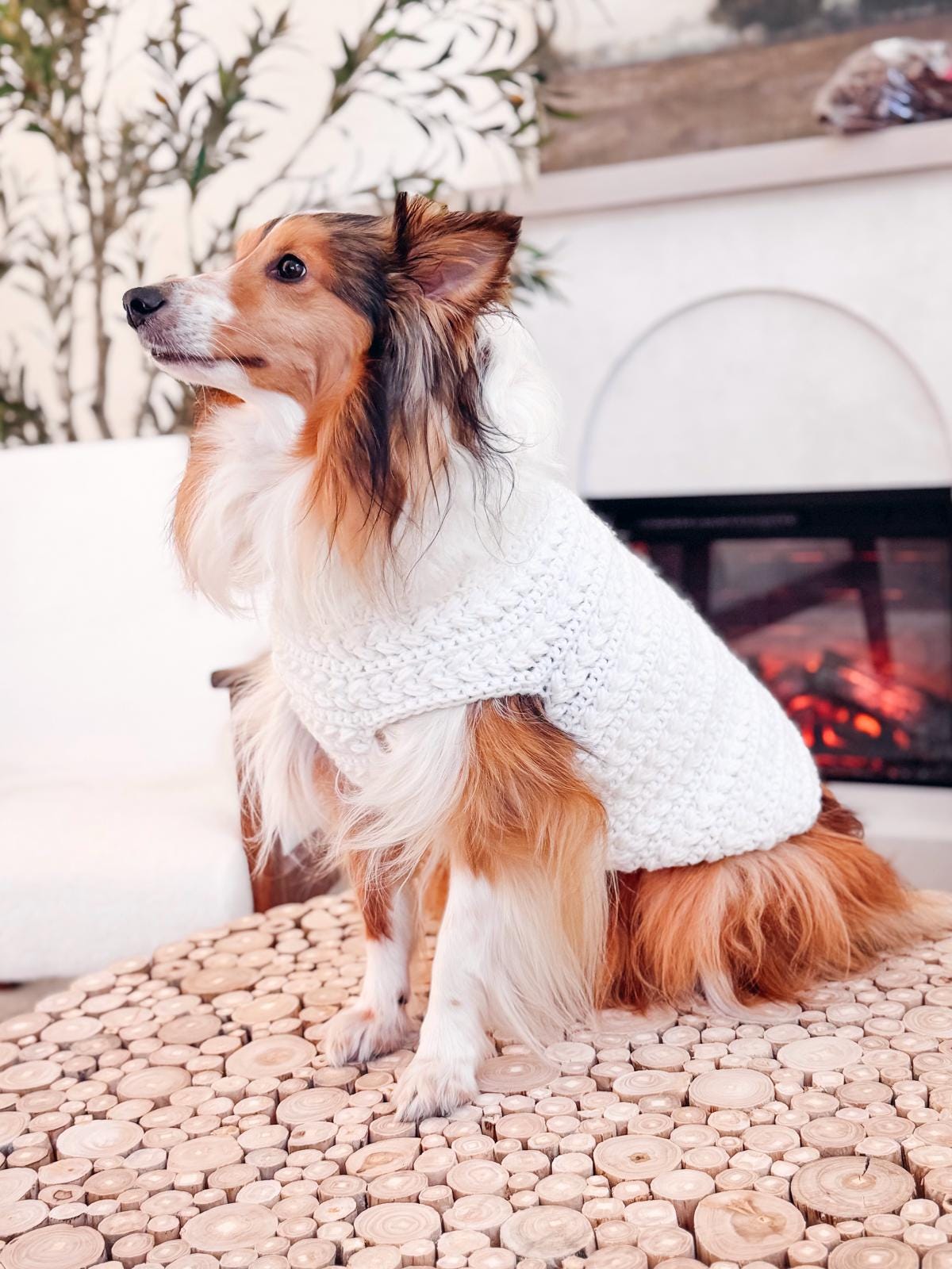 A Shetland Sheepdog wearing a white crocheted sweater sits on a round wooden table in front of a fireplace, beside a handmade gift and some vintage clothes, creating a cozy and charming scene.