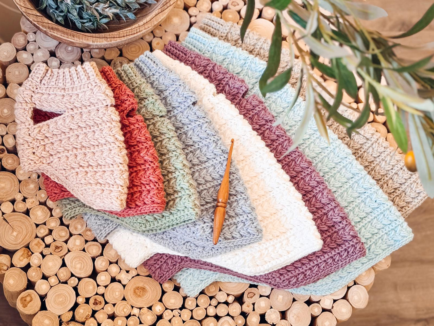 Several crocheted dog sweaters in pastel colors are neatly arranged in a fan shape on a round wooden table, with a crochet hook placed on top—perfect as a handmade gift for someone special.