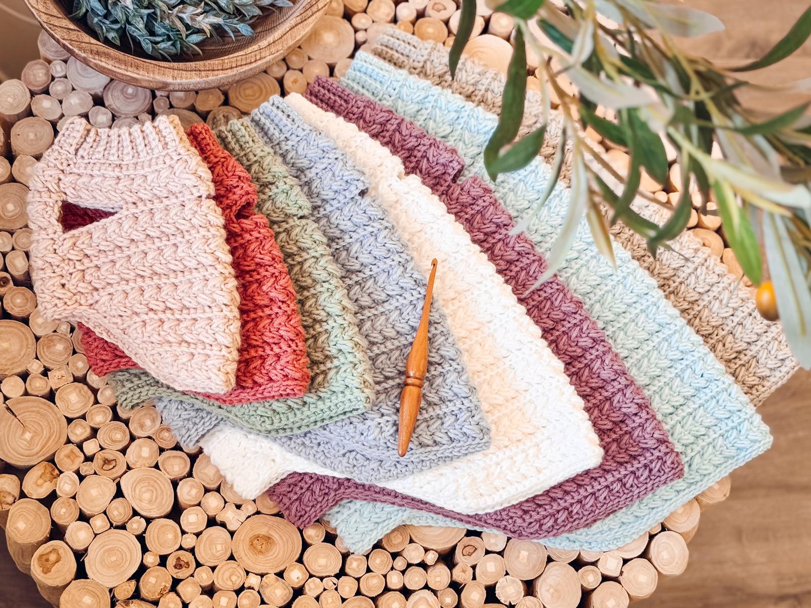 Several crocheted dog sweaters in pastel colors are neatly arranged in a fan shape on a round wooden table, with a crochet hook placed on top—perfect as a handmade gift for someone special.