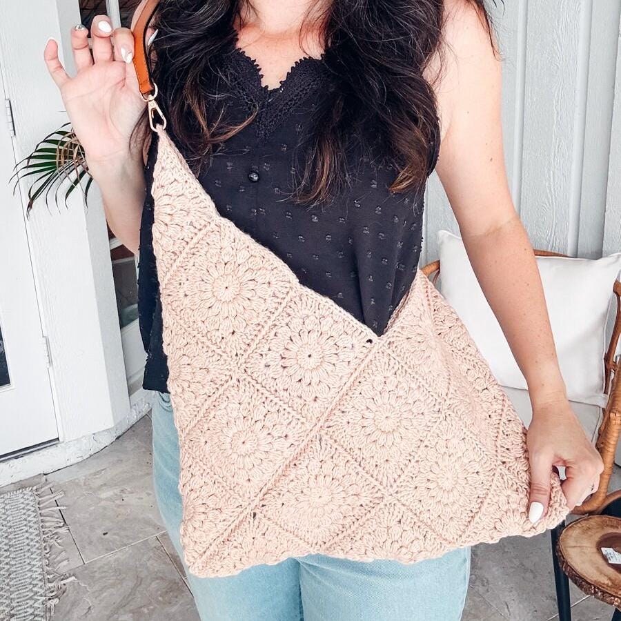 A woman in a black top and jeans holds a beige, crocheted handbag, showcasing her flair for vintage clothes. She is standing indoors near a white door.