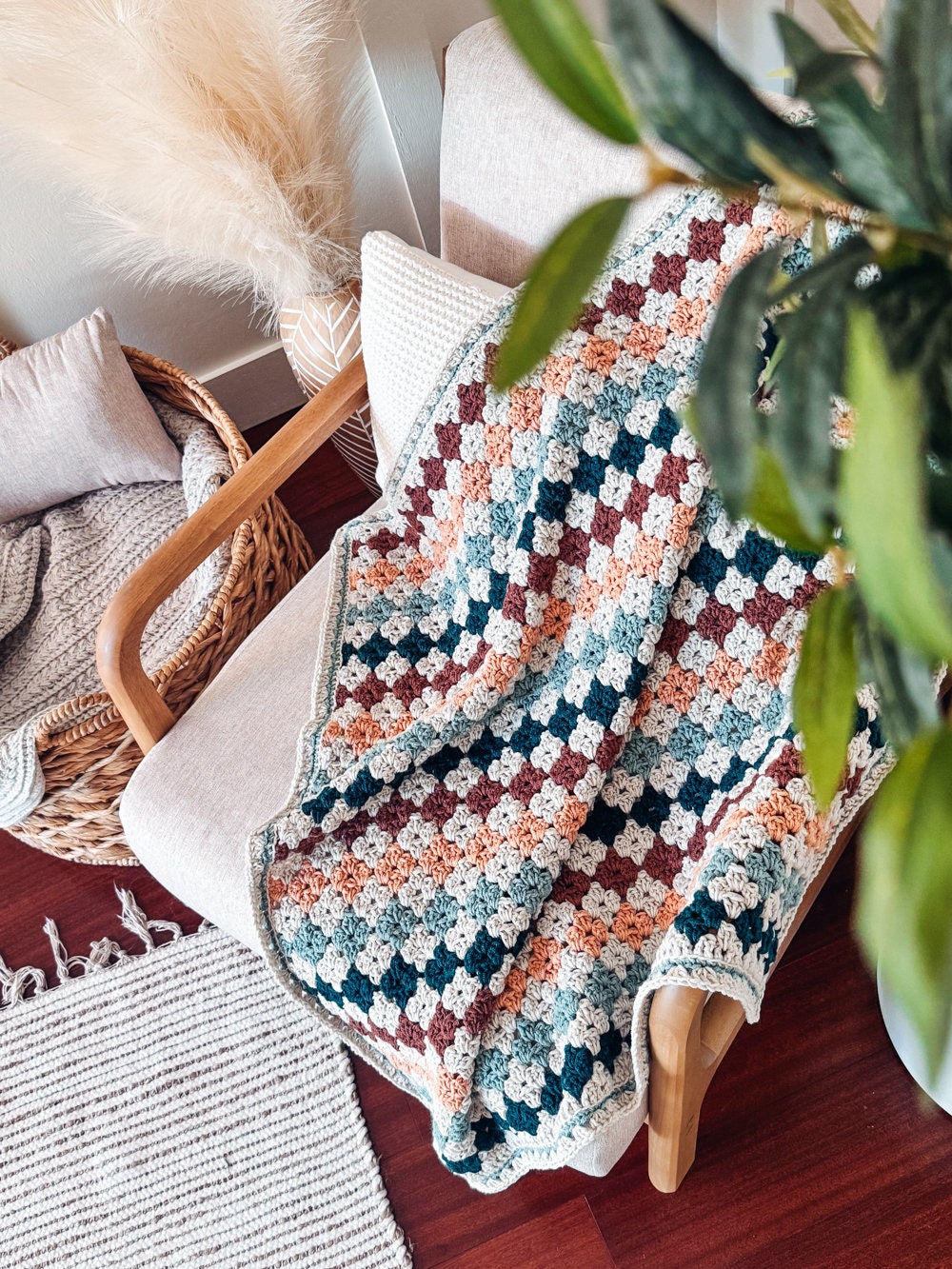 May include: A crocheted blanket with a checkered pattern in shades of brown, green, blue, and orange. The blanket is draped over a chair with a white cushion and a wooden armrest. A potted plant with green leaves is in the background.
