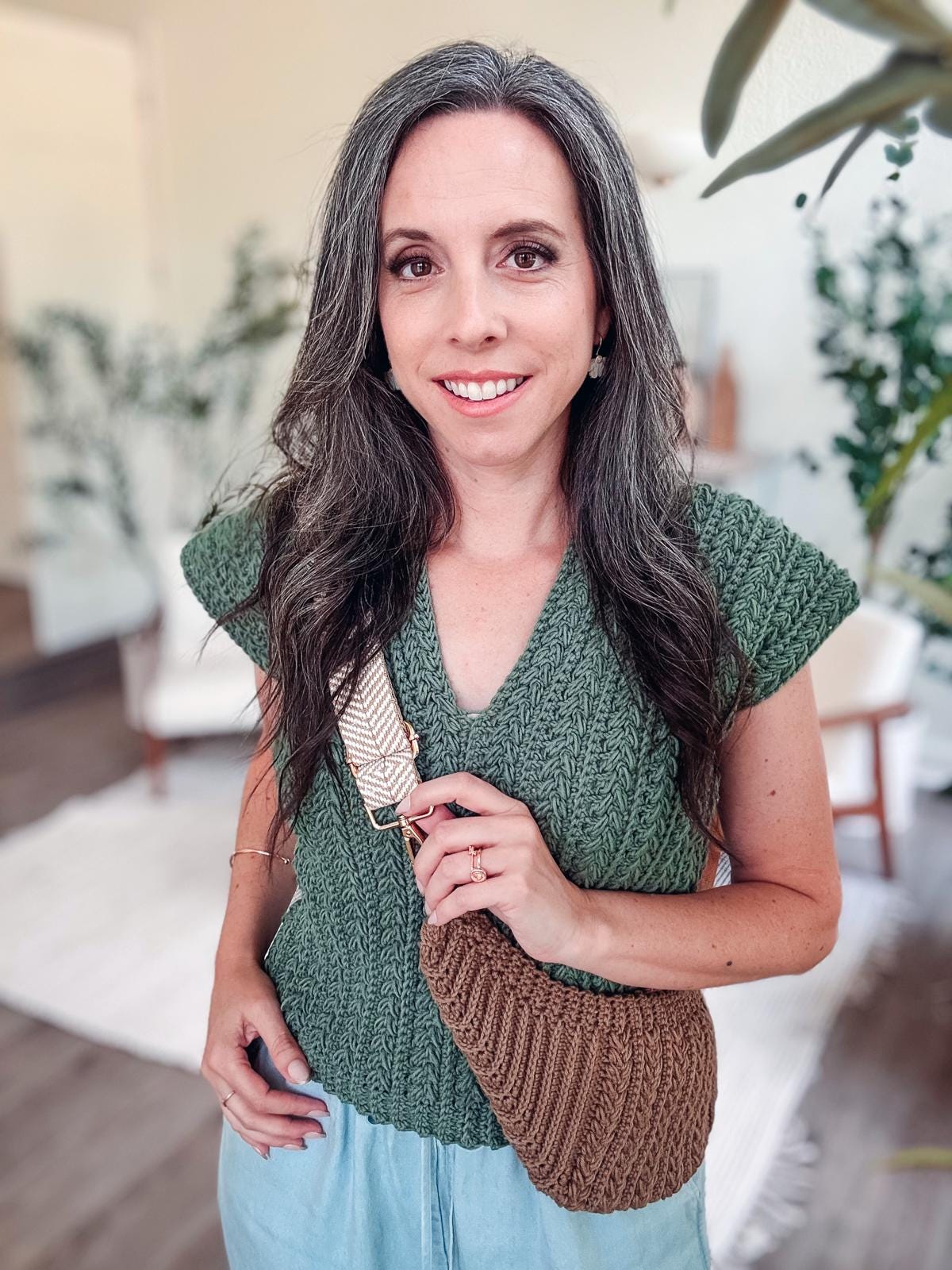 A person with long hair stands indoors, wearing a green crochet top and holding a brown crocheted bag, complemented by unique jewellery that adds an elegant touch.