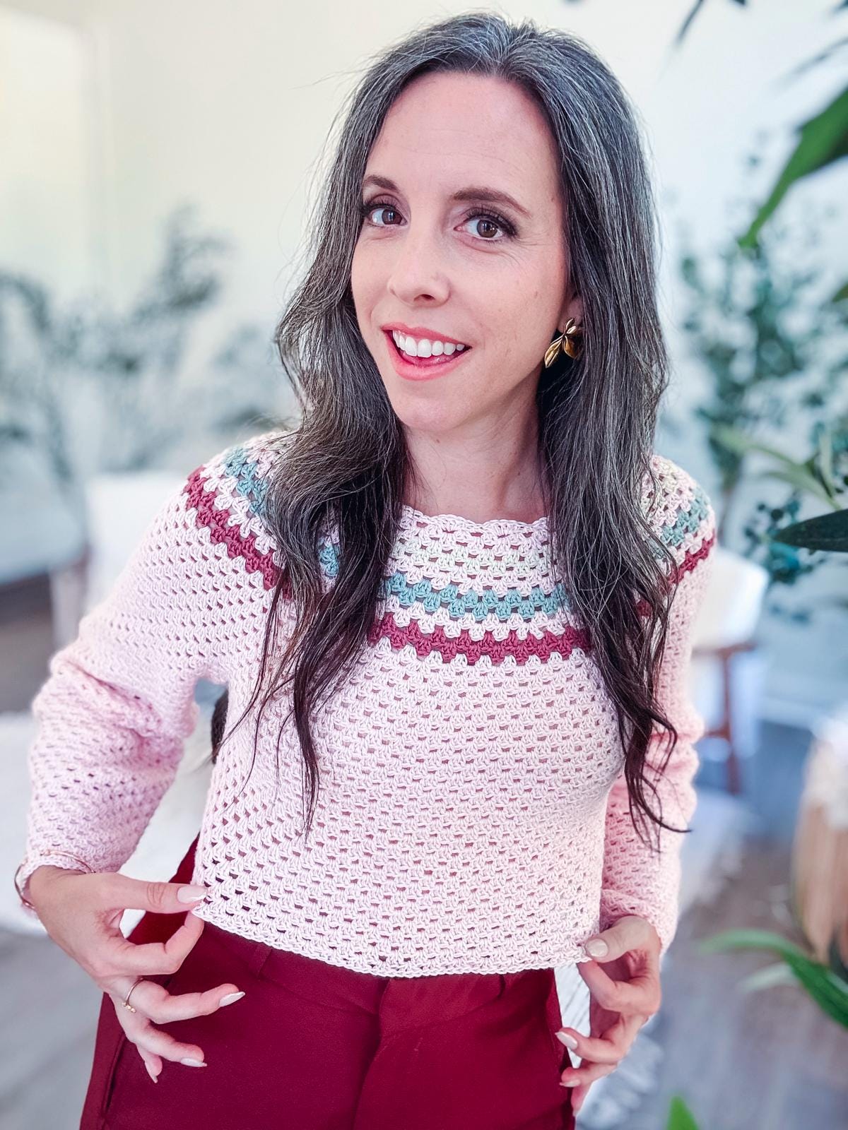 A woman with long hair smiles while wearing a pink crocheted sweater with colorful detailing and maroon pants. She stands in a room with plants, accessorizing her look with unique jewelry that perfectly complements her shorts.
