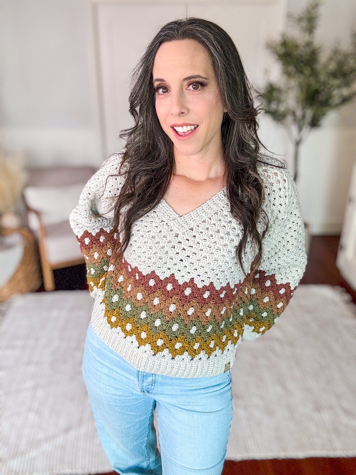 A person with long, dark hair, wearing a multicolored crochet sweater and light blue jeans, stands in a room with hardwood floors, a woven chair, unique jewelry, and a leafy plant in the background.