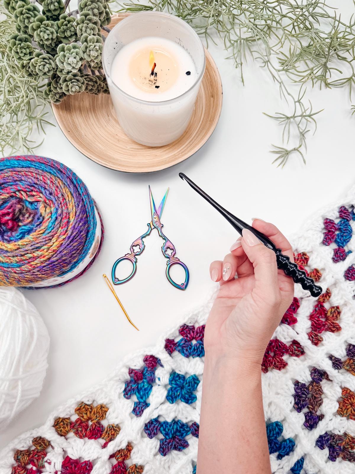 A hand holds a black crochet hook above a multicolored yarn skein, scissors, a yarn needle, and a candle on a table with a crocheted blanket and plants—perfect for crafting a handmade gift.