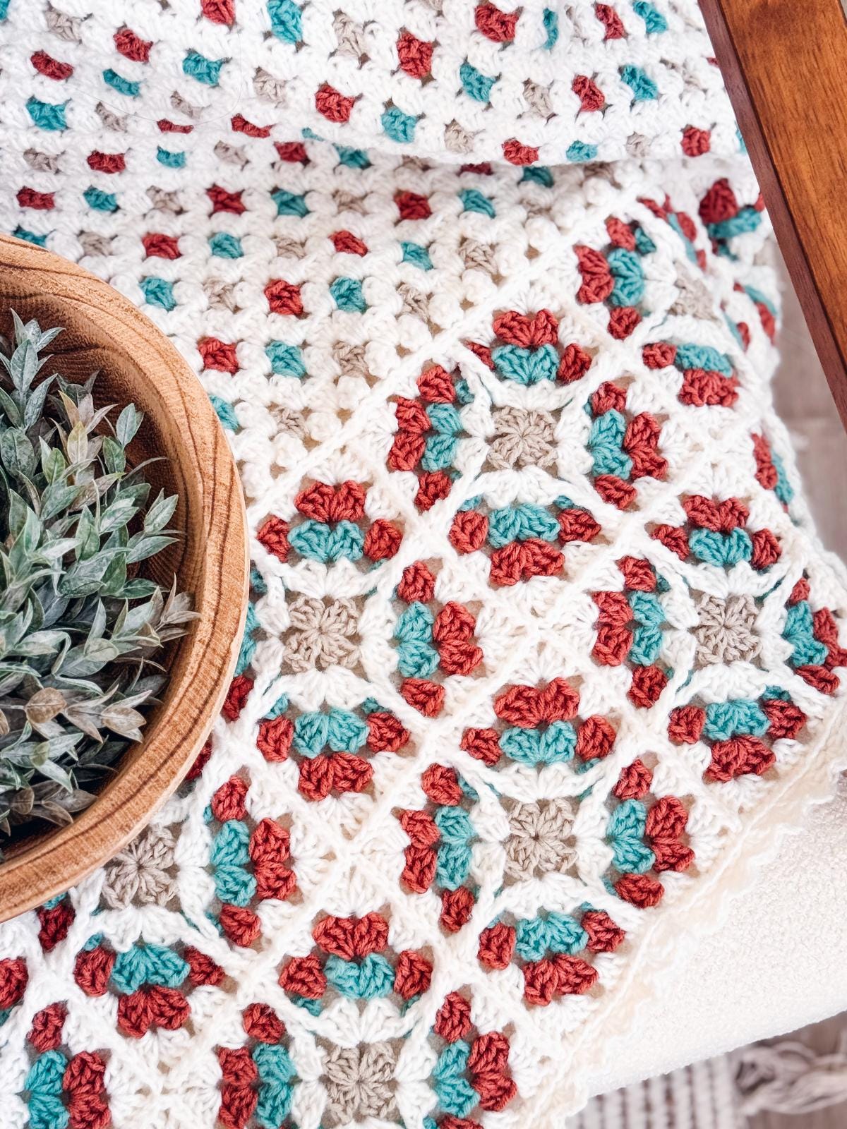 A close-up of a crocheted blanket featuring a pattern in white, red, teal, and beige yarn, next to a wooden bowl holding a succulent plant—perfect as a handmade gift for those who love cozy decor or vintage clothes.
