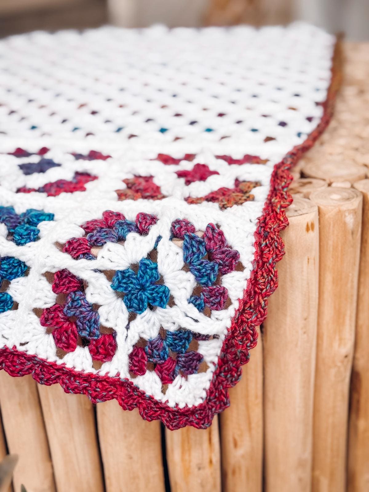 A crochet blanket with a white base and multicolored granny squares, edged in red, draped over a wooden surface—a perfect handmade gift.