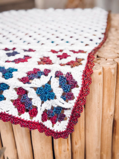 Corner of white crochet granny square blanket with blue and red motifs and a red edge on a log side table