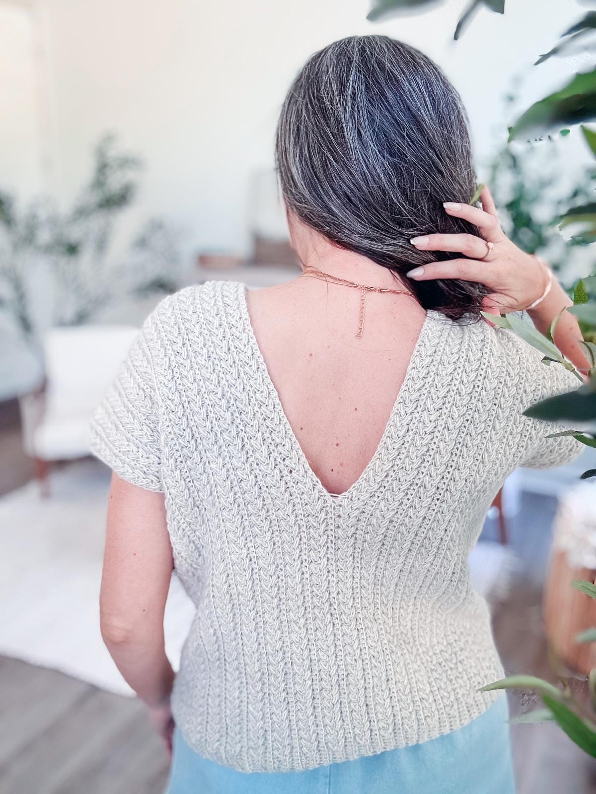 A person with gray hair is wearing a textured crochet top with a deep V-back, standing indoors amidst plants, accentuated by unique jewellery.