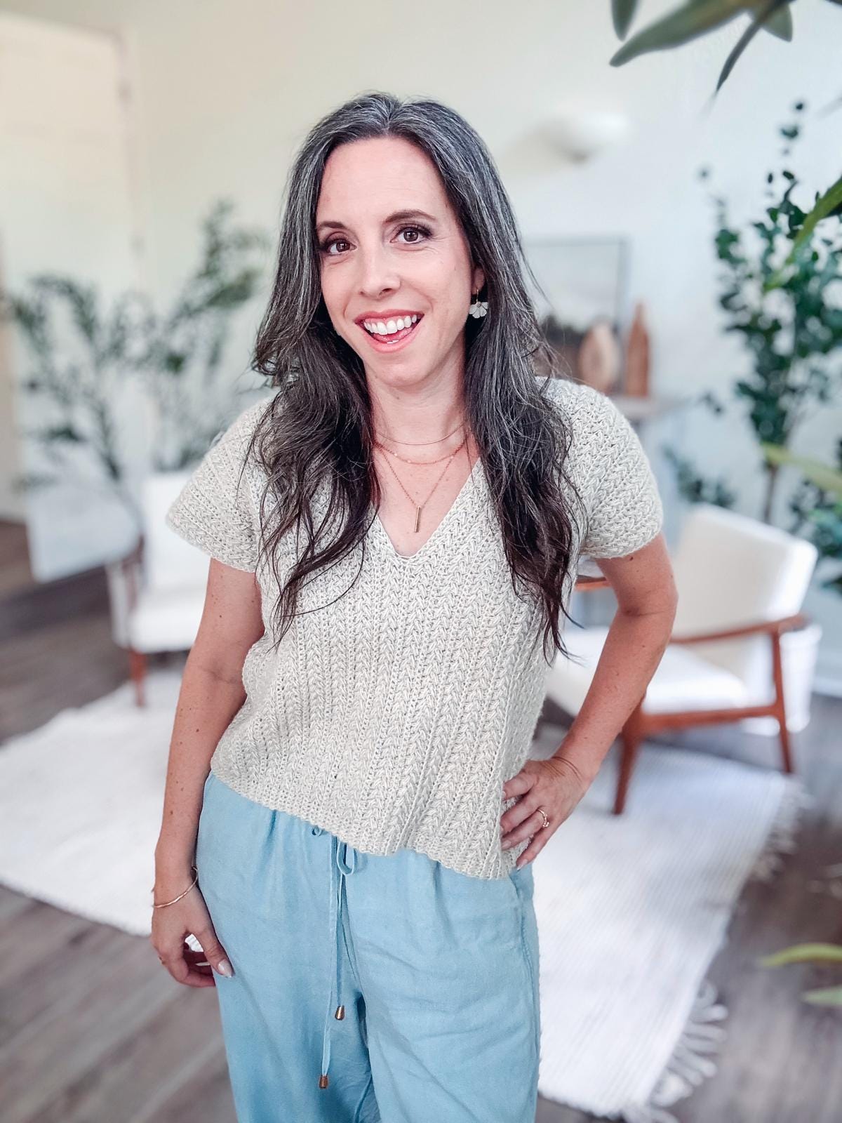 A woman with long hair stands in a room, adorned in a crochet short-sleeve top and blue pants. Unique jewellery catches the eye, adding charm. In the background, lush plants and vintage chairs create an inviting atmosphere.