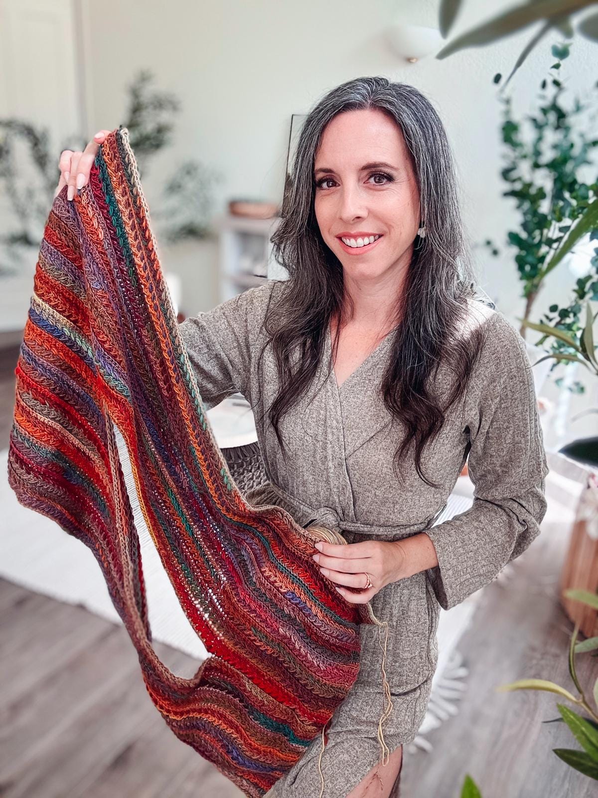 A woman in a gray outfit holds a colorful, striped crocheted piece in a room with plants, surrounded by vintage clothes.