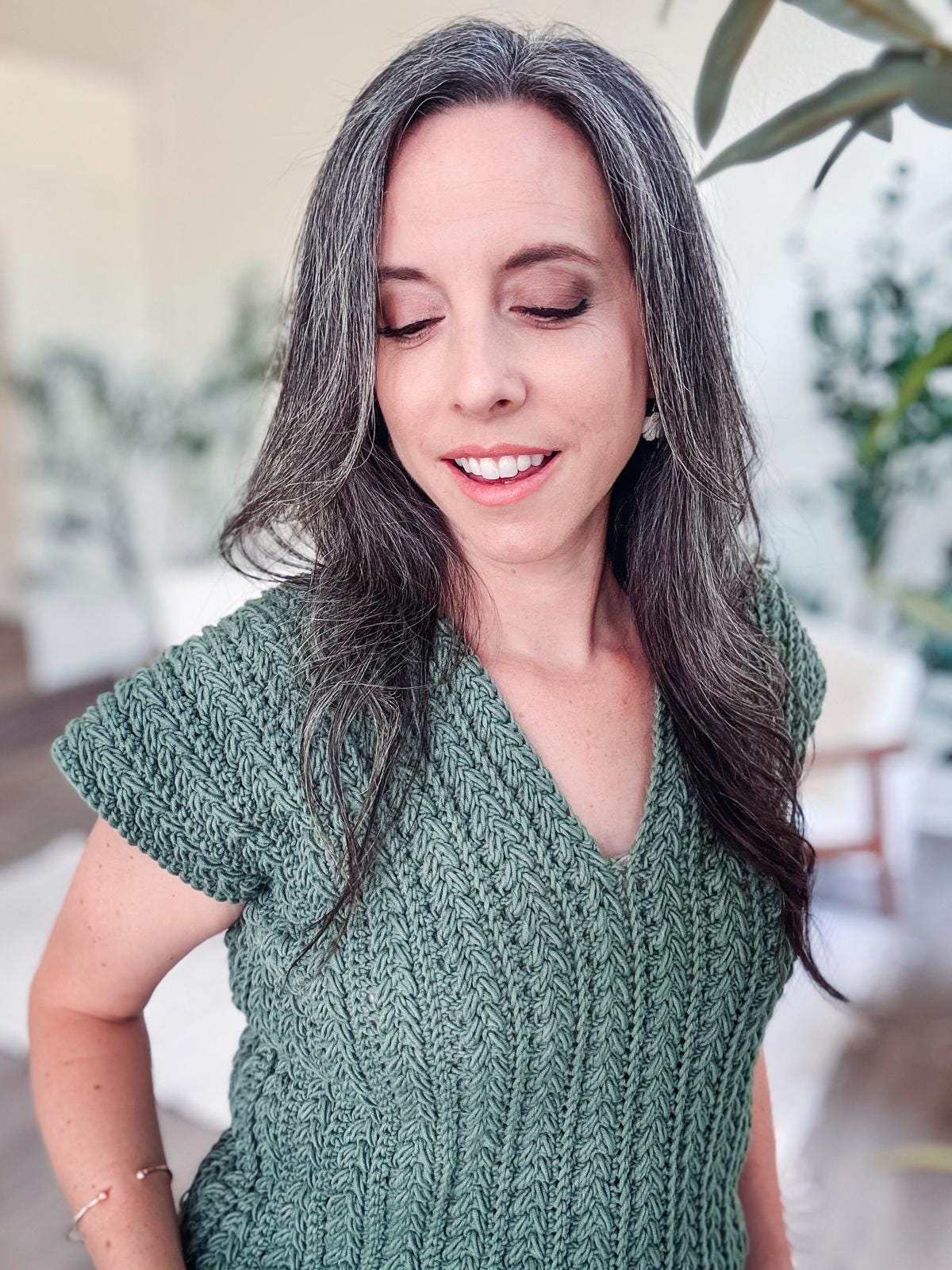 Person with long, gray hair wearing a textured green crochet top, standing indoors with eyes closed, smiling gently, adorned with unique jewellery that adds a touch of elegance.