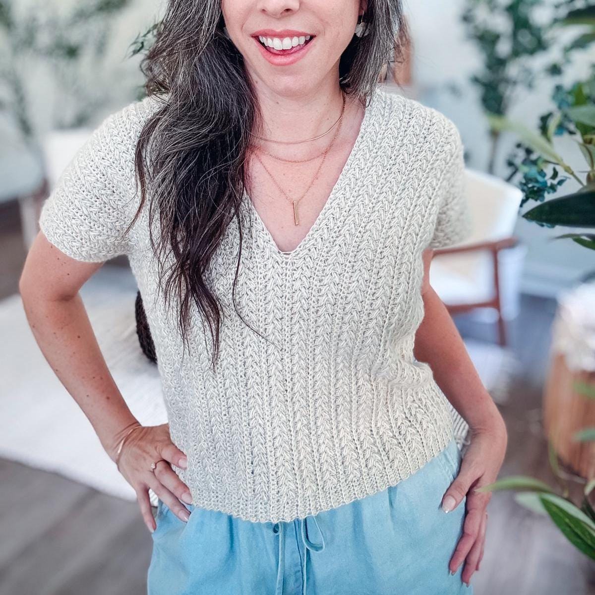 A woman with long gray hair wearing a textured crochet V-neck sweater and blue pants stands indoors, surrounded by greenery. Her ensemble is complemented by unique jewellery that adds an artistic flair to her vintage-inspired look.