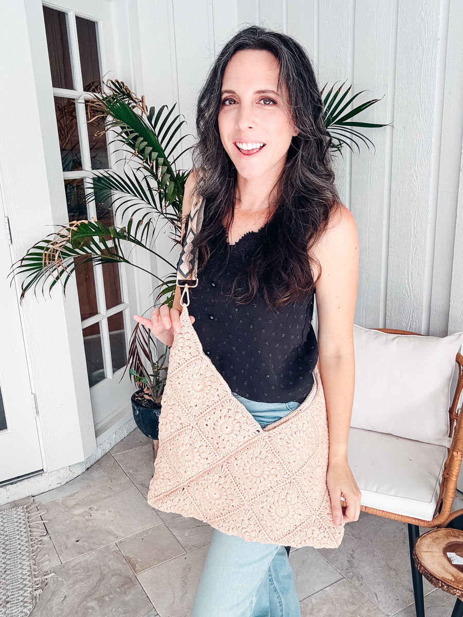 Woman standing by a plant indoors, wearing a black sleeveless top, light blue jeans, and holding a crochet bag. She admires her handmade gift—a piece of unique jewellery that complements her vintage clothes perfectly.