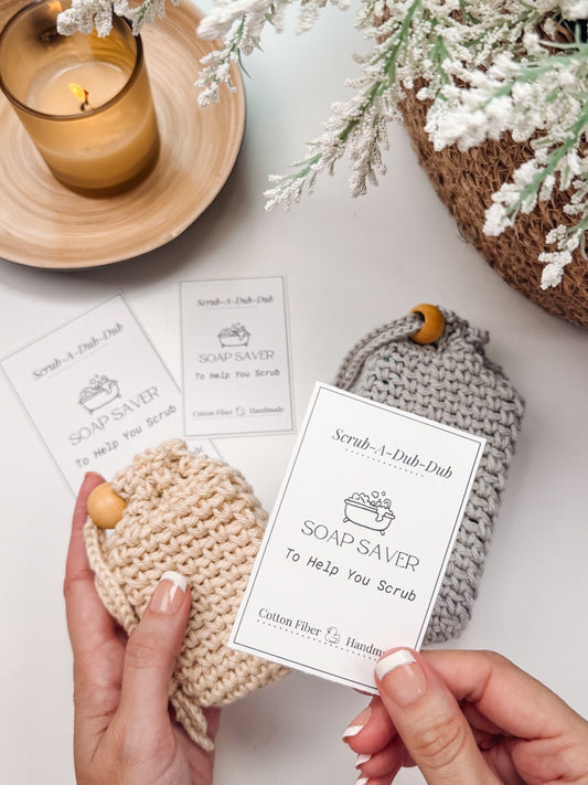 A person holds a beige 'Soap Saver' pouch with a white product card; a grey pouch and another card rest on the surface. A lit candle and a plant are in the background, creating an inviting atmosphere perfect for showcasing handmade gifts.