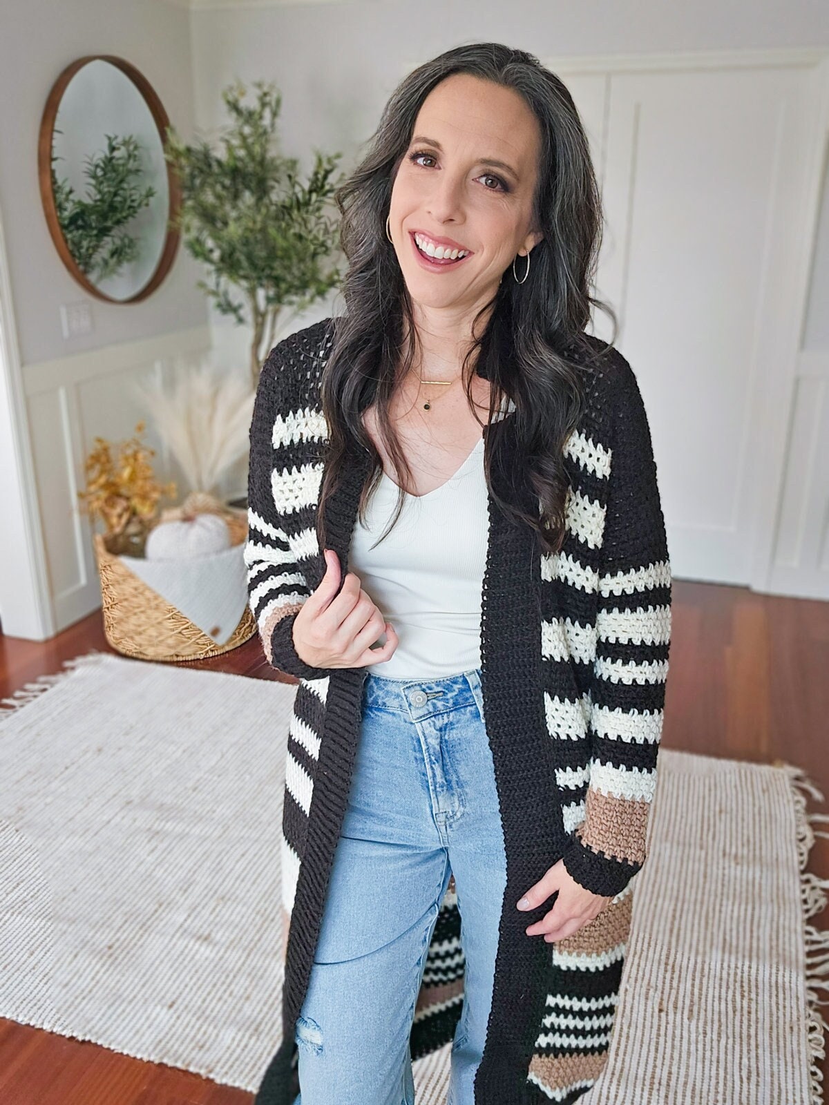 A woman with long dark hair stands in a room with white walls and hardwood floors, wearing a striped cardigan and blue jeans. She smiles at the camera, her hand holding her cardigan, the other down. Her unique jewellery completes this charming look.
