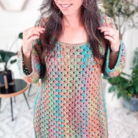 A person is wearing a colorful granny stitch crochet dress, standing in a room with geometric wall decor and plants in the background, accessorized with unique jewelry.