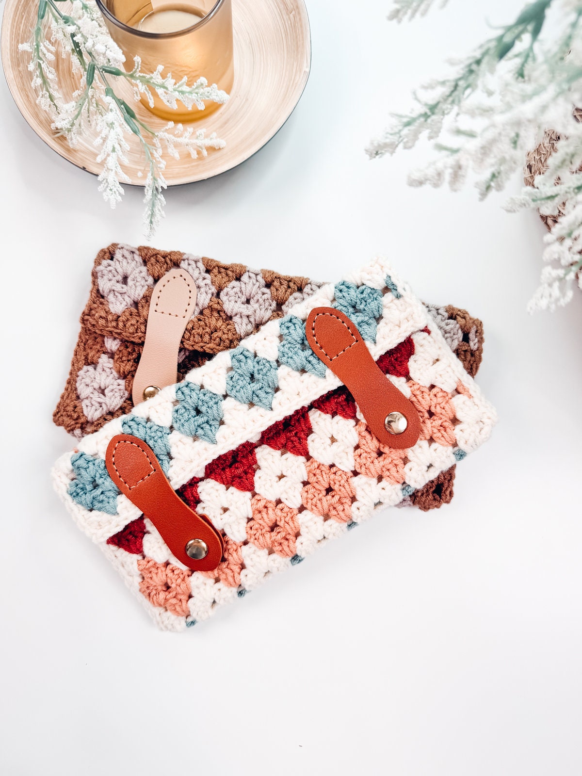 Two crocheted pouches with leather straps are placed on a white surface, accompanied by a glass of liquid and some decorative greenery, making for a charming handmade gift.